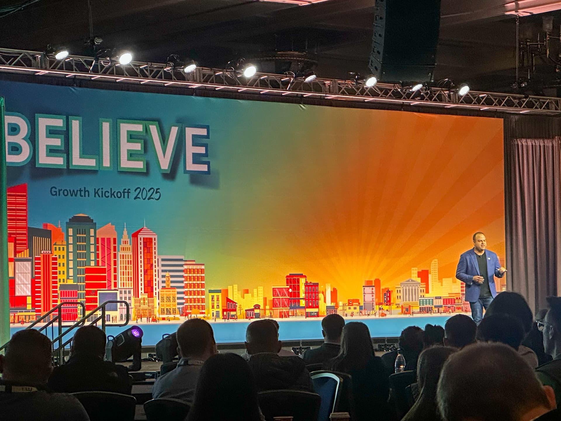 David on stage at the BELIEVE Growth Kickoff 2025, speaking to a full audience.
